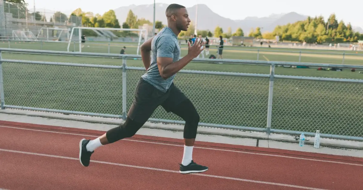 An individual with a pixelated face is sprinting on a red running track, wearing a gray t-shirt, black shorts, and white socks with black sneakers. The background features a soccer goal and mountains in the distance under a clear sky.