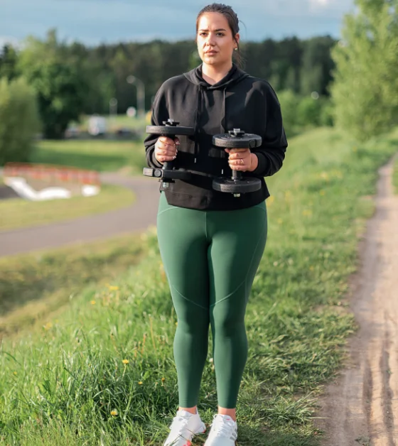 A woman in a black hoodie and green leggings stands on a grassy path, holding two dumbbells. She is outdoors with trees and a field visible in the background.