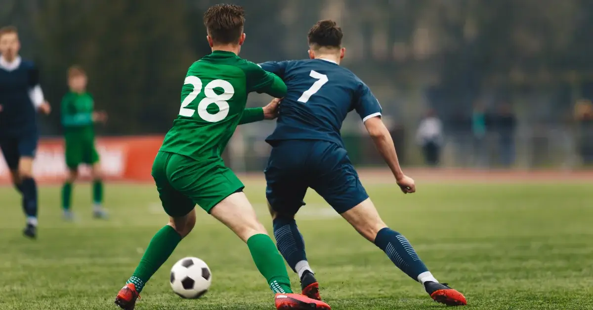 Two soccer players in mid-action on the field, one wearing a green jersey with the number 28 and the other in a navy blue jersey with the number 7, both intensely focused on a soccer ball at their feet.