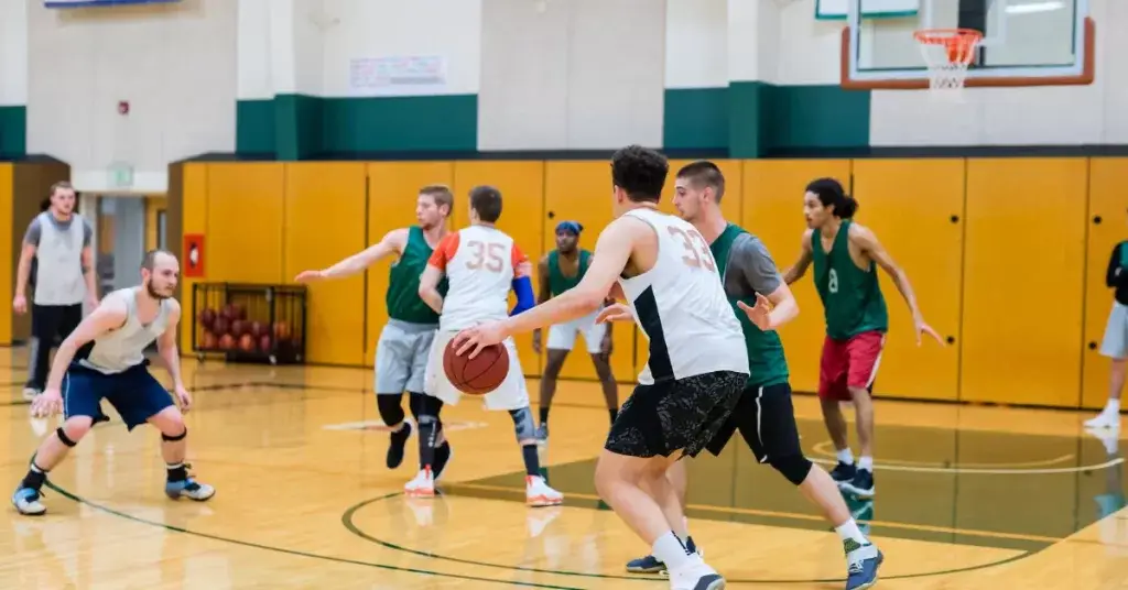 An indoor basketball game in progress with players from two teams. One player is dribbling the ball while others are in various positions around the court, some defending and others preparing to receive the ball.