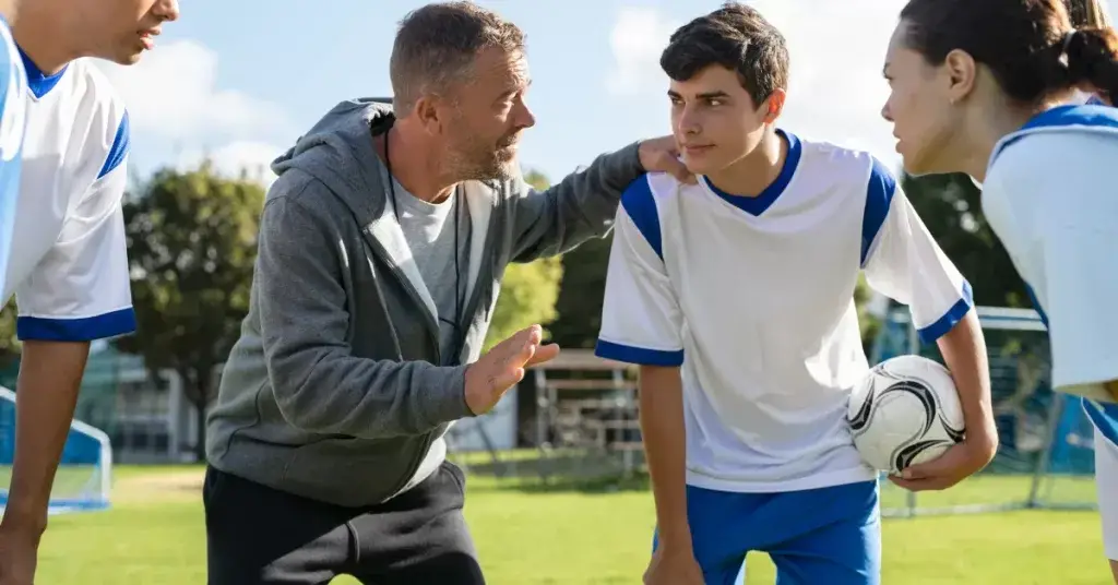 A group of soccer players on a field, with two individuals in the foreground discussing strategy while holding a soccer ball, and three teammates watching in the background.