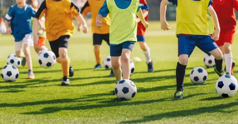 A group of children in colorful sportswear playing soccer on a grassy field, with multiple soccer balls in play.